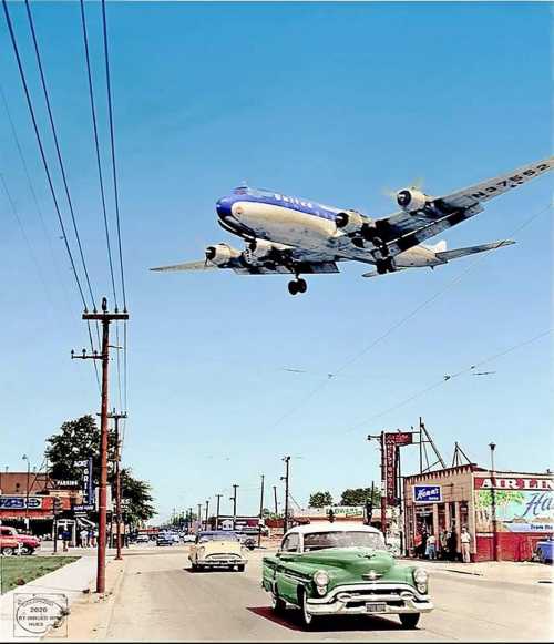 A vintage scene with a green car and an airplane flying overhead in a clear blue sky, showcasing a bustling street.