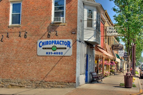 A brick building with a chiropractor sign and nearby shops, featuring flowers and outdoor seating along the sidewalk.