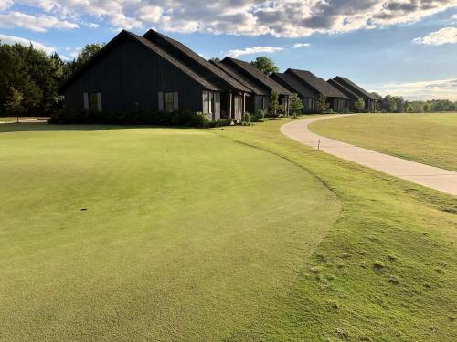 A view of a golf course with green grass and several dark-roofed buildings in the background under a cloudy sky.