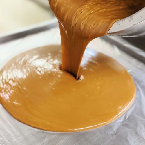 A smooth, caramel-colored mixture is being poured onto a flat surface, creating a glossy layer.