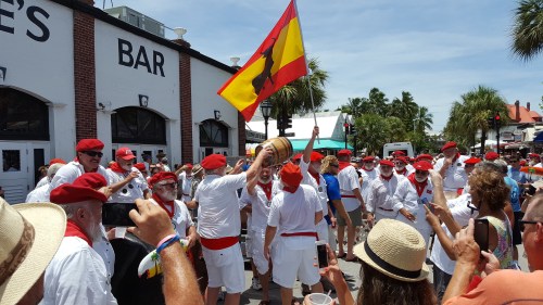 A lively street scene with people in white outfits and red accessories celebrating, waving a flag, and enjoying the festivities.