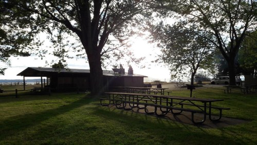 A park scene with picnic tables, a shelter, and sunlight filtering through trees near a body of water.