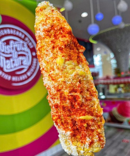 A close-up of a grilled corn on the cob, coated with cheese and spices, hanging against a colorful background.
