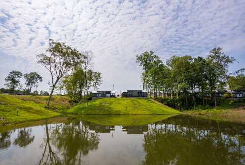 A serene landscape featuring a pond reflecting green hills and trees, with modern buildings in the background under a cloudy sky.