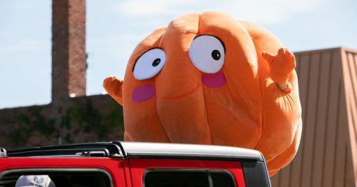 A large, cheerful orange character with big eyes and a smiling face peeks out from a red vehicle.
