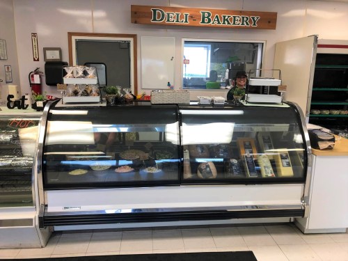 A deli-bakery display case filled with various food items, with a staff member visible behind the counter.