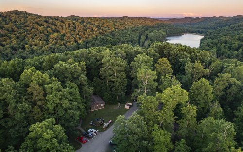 Aerial view of a wooded area with a small lake, surrounded by lush trees and a few parked cars near a cabin.