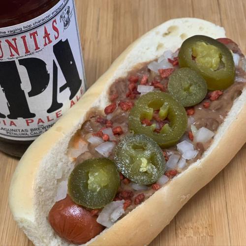 A hot dog in a bun topped with refried beans, onions, jalapeños, and bacon bits, alongside a bottle of IPA beer.