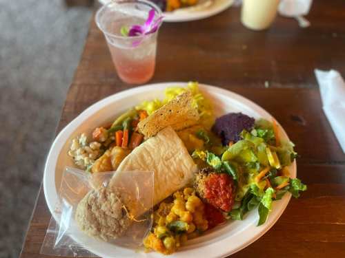 A plate of colorful food including salad, vegetables, a tortilla, and a cookie, with a drink in the background.