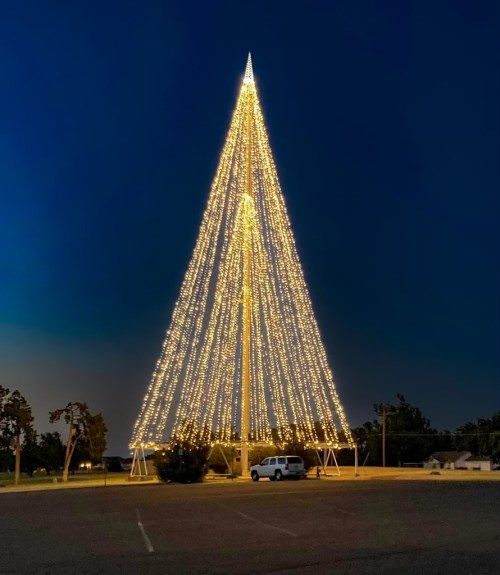 A tall, illuminated Christmas tree made of lights stands in a dark parking lot at night.