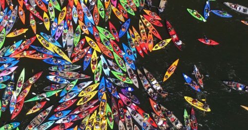 Aerial view of a vibrant gathering of colorful kayaks and canoes on a dark water surface.