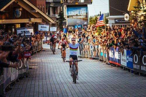 A cyclist celebrates victory while crossing the finish line, surrounded by cheering fans and waving flags.