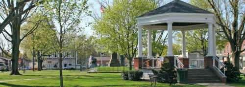 A sunny park scene featuring a white gazebo surrounded by green trees and grass, with buildings in the background.