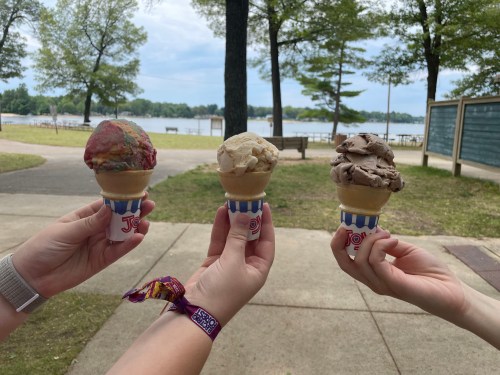 Three hands hold ice cream cones of different flavors against a scenic lakeside background.