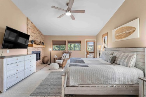 A cozy bedroom featuring a large bed, TV, fireplace, and natural light from windows, with neutral decor and a rug.