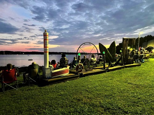 A crowd sits on the grass by a lake at sunset, enjoying the view with boats in the water and colorful clouds above.