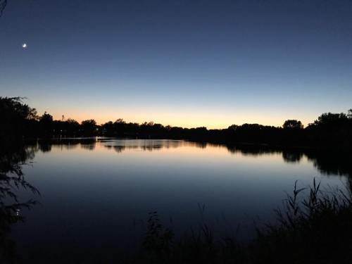 A serene lake at dusk, reflecting the twilight sky and silhouettes of trees along the shore. A crescent moon is visible.