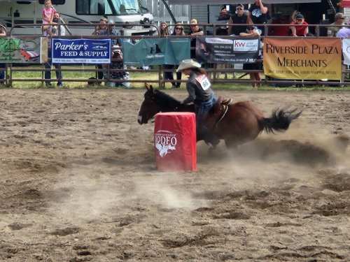 A rider on a horse races around a barrel in a rodeo arena, with spectators and banners in the background.