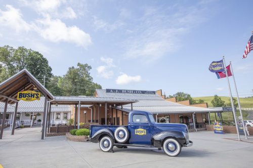 A vintage blue truck parked in front of the A.J. Bush Co. building, with flags and greenery in the background.