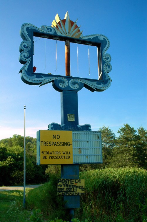 An empty, vintage sign structure with a "No Trespassing" notice at the base, surrounded by greenery.