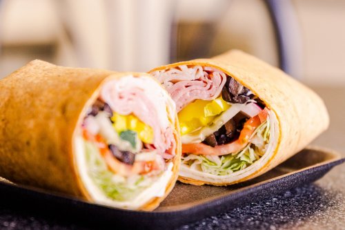 A close-up of a sliced wrap filled with meats, lettuce, tomatoes, and pickles on a dark plate.