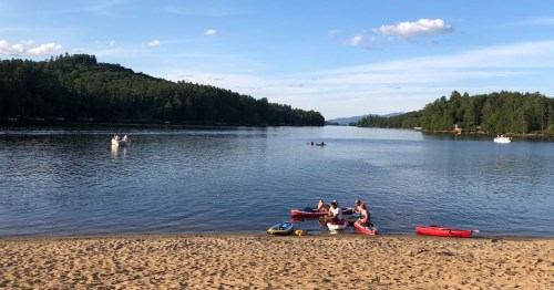 A serene lake scene with kayakers on the water, surrounded by lush green trees and a sandy beach.
