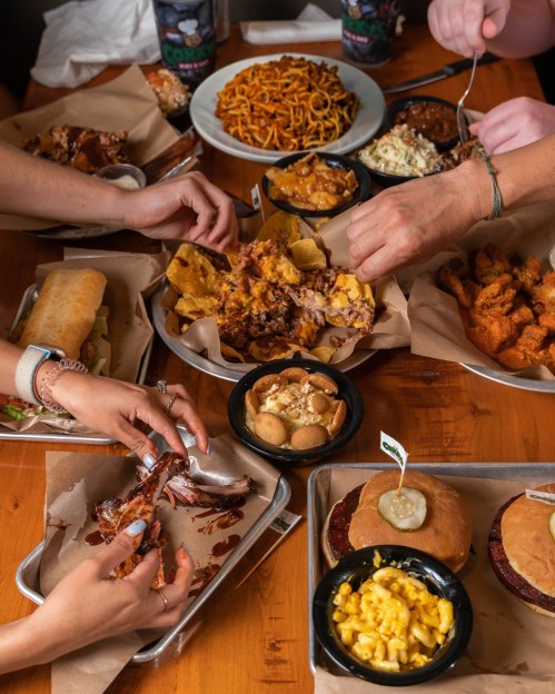 A table filled with various dishes, including ribs, nachos, sandwiches, and sides, with hands reaching for the food.