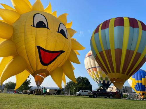 Colorful hot air balloons, including a smiling sun design, are inflated on a grassy field under a clear blue sky.
