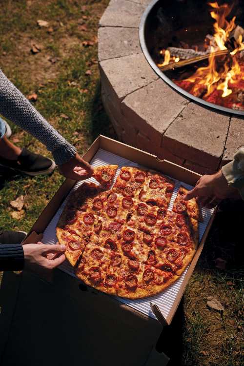 Two people are sharing a pepperoni pizza near a fire pit, surrounded by grass and autumn leaves.