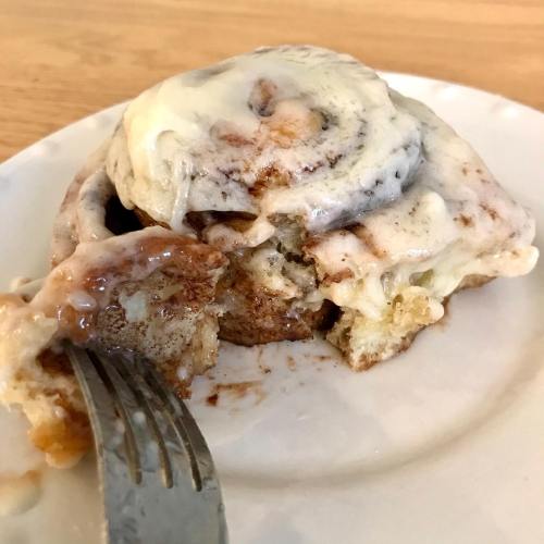 A partially eaten cinnamon roll with cream cheese frosting on a white plate, with a fork resting beside it.