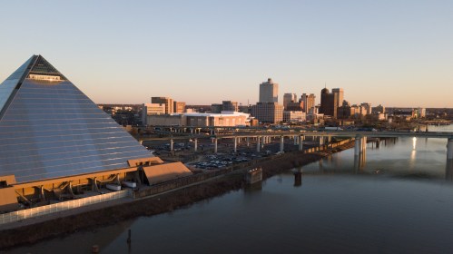 Aerial view of a pyramid-shaped building beside a river, with a city skyline in the background during sunset.