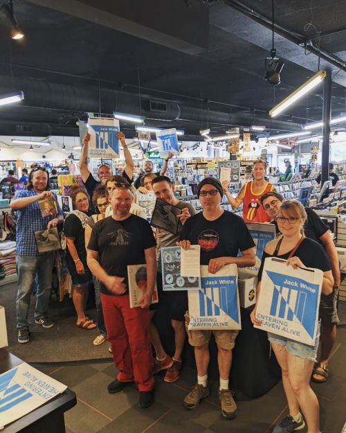 A group of people in a store hold up Jack White merchandise and posters, smiling and celebrating together.