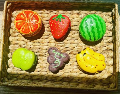 A woven basket filled with colorful macarons shaped like fruits: orange, strawberry, watermelon, green apple, grapes, and bananas.