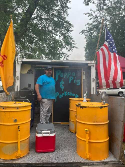 A man stands in front of a food trailer named "Poppa Pig & Porkchop," with yellow barrels and flags nearby.