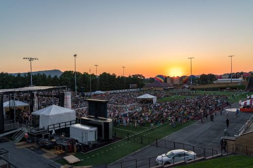 A large crowd gathers at an outdoor event as the sun sets, with colorful hot air balloons in the background.