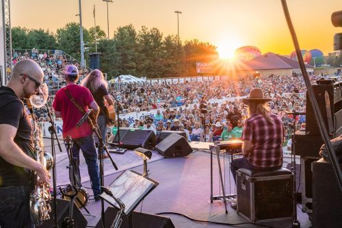 A band performs on stage at sunset, with a large crowd enjoying the concert in the background.