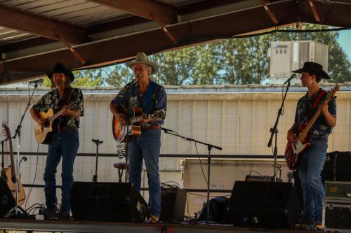 Three musicians perform on stage, playing guitars and singing under a metal roof with instruments and equipment nearby.