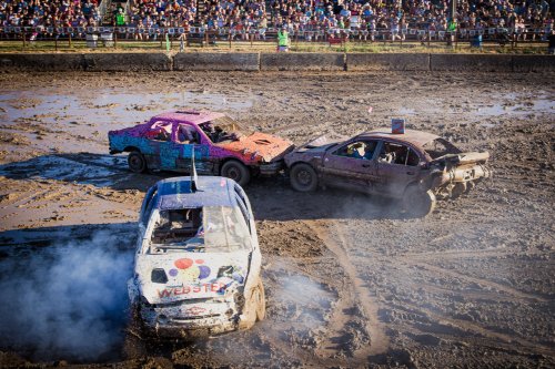 Three cars collide in a muddy arena during a demolition derby, with a crowd watching in the background.