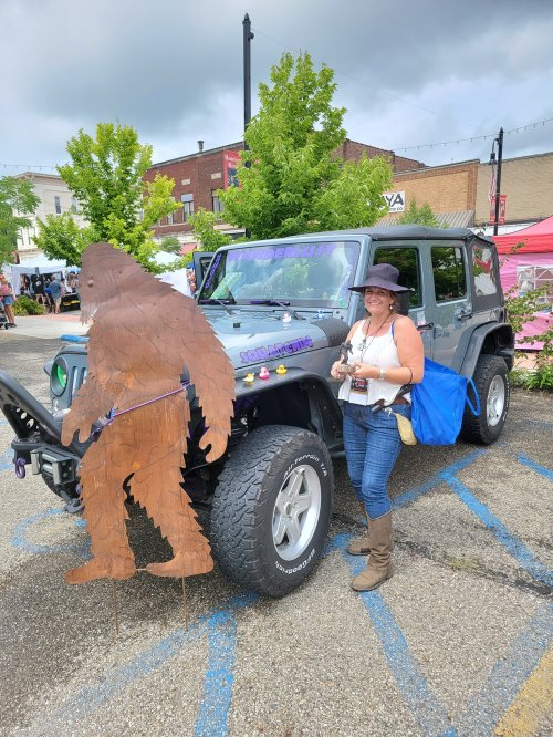A woman stands next to a decorated Jeep and a cutout of Bigfoot at an outdoor event.