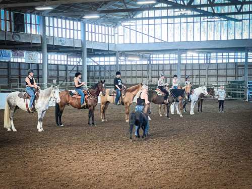A group of riders on horseback in an indoor arena, with a person on the ground interacting with a horse.