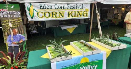 A festival booth displaying corn with a sign reading "Eden Corn Festival" and "Corn King," featuring fresh corn cobs.
