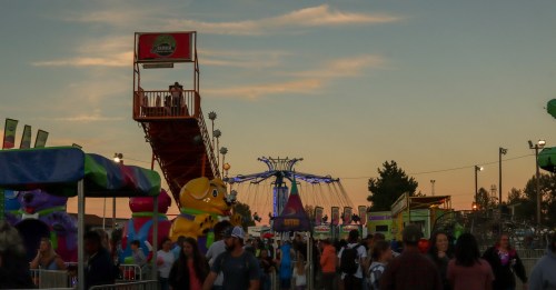 A lively carnival scene at sunset, featuring rides, colorful booths, and a crowd of people enjoying the festivities.