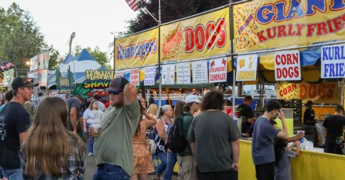 A bustling fair scene with people walking past food stalls selling corn dogs and curly fries under colorful banners.