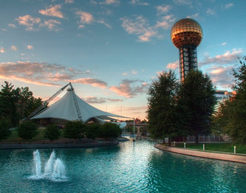A scenic view of a park with a golden observation tower, a tent structure, and a pond with fountains under a colorful sky.