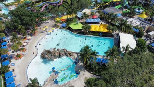 Aerial view of a vibrant water park with pools, slides, and colorful cabanas surrounded by palm trees.