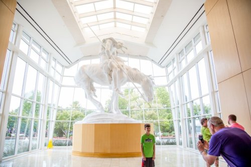 A large white statue of a horse and rider in a bright, modern atrium with visitors taking photos.