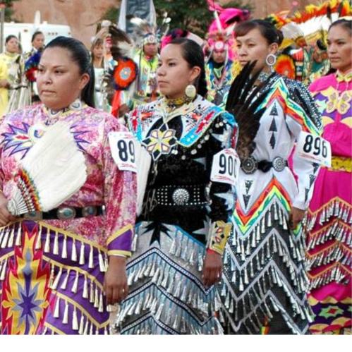 A group of Indigenous women in colorful, traditional regalia, standing together at a cultural event.