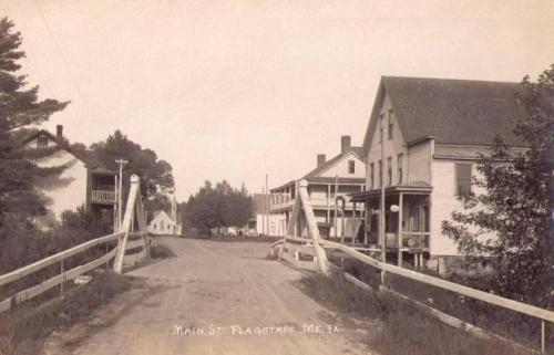 Black and white photo of a quiet street in Flagstaff, Maine, featuring wooden houses and a bridge.