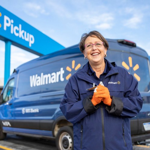 A smiling Walmart employee in a blue jacket stands in front of a Walmart pickup van, holding a pen and wearing orange gloves.