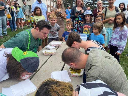 Participants compete in a food-eating contest, leaning over tables with burgers, surrounded by an enthusiastic crowd.